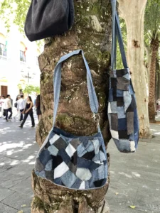 Collection de sacs Nat_Collection disposés sur un tronc de palmier moussu dans un environnement urbain méditerranéen : Sac Signature noir, Patch Bag et Tote Bag en patchwork de jean recyclé, illustrant la gamme complète "On the Move"