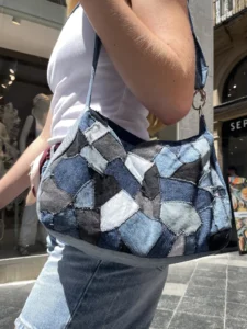 Gros plan sur un sac à bandoulière en patchwork de jean recyclé porté par une femme dans un environnement commercial urbain, montrant les détails de couture et la variété des tons de denim de la collection Nat_Collection