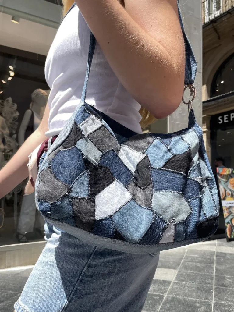 Gros plan sur un sac à bandoulière en patchwork de jean recyclé porté par une femme dans un environnement commercial urbain, montrant les détails de couture et la variété des tons de denim de la collection Nat_Collection