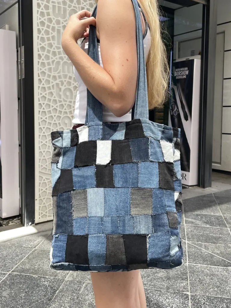 Texte alternatif : Femme blonde portant le Tote Bag en patchwork de jean recyclé de Nat_Collection dans un environnement urbain commercial, montrant le grand format et les motifs géométriques en damier du sac éco-responsable