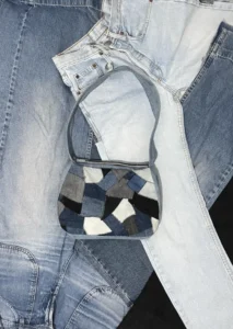 Sac à bandoulière Nat_Collection en patchwork de jean recyclé posé sur un arrangement de jeans de différentes nuances, illustrant le processus d'upcycling et la transformation créative des matériaux de seconde main en accessoires uniques